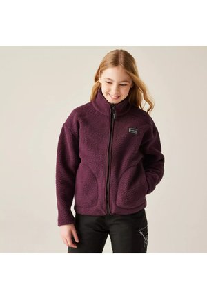 Jonge persoon met een paarse fleecejas met ritssluiting en hoge kraag en zwarte broek, staand met één hand in de zak, glimlachend naar beneden.