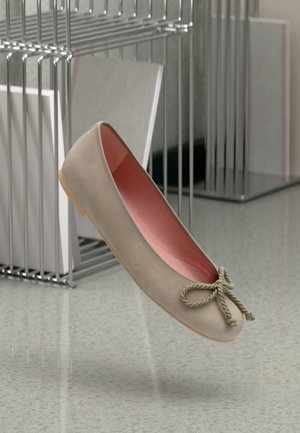 Chaussure ballerine beige avec un nœud en corde tordue, doublure intérieure rose et semelle marron clair, flottant au-dessus d’un sol réfléchissant près de rayonnages métalliques.