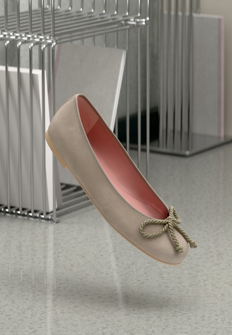 Chaussure ballerine beige avec un nœud en corde tordue, doublure intérieure rose et semelle marron clair, flottant au-dessus d’un sol réfléchissant près de rayonnages métalliques.