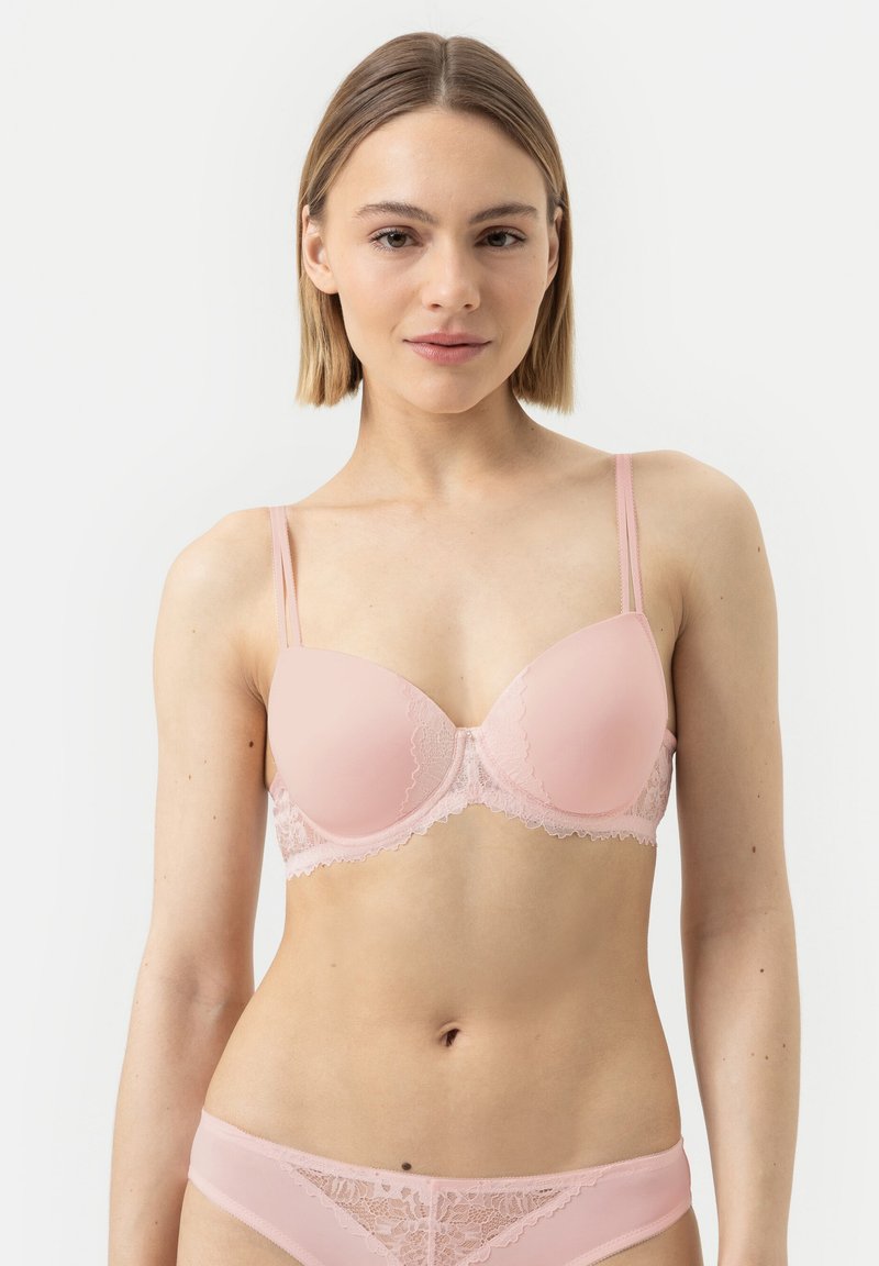 Vrouw in een bijpassende lichtroze kanten beha en slip tegen een effen witte achtergrond, recht vooruit kijkend.