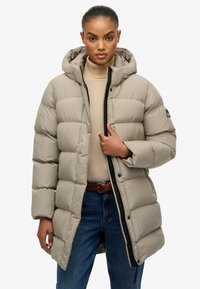 Chaqueta acolchada beige con capucha, que presenta secciones acolchadas horizontales, cremallera negra y detalles de bolsillo. Lleva puesta una camiseta de cuello alto beige.