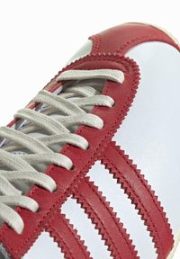 Röd och vit lädersneaker med texturerade röda ränder, vita snören och en sydd detalj, som har en slät ovandel och gummisula.