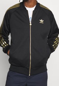 Svart zip-jacka med guldaccenter. Har ribbade ärmslut och nederkant, polyester material, och tre guldstripes på ärmarna. Adidas-logga på bröstet.