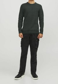 Barn i en mörkgrön texturerad tröja, svarta cargo-byxor med sidofickor och svarta sneakers med vita sulor, står mot en enfärgad bakgrund.