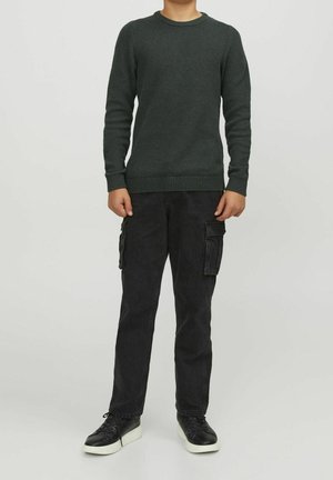 Barn i en mörkgrön texturerad tröja, svarta cargo-byxor med sidofickor och svarta sneakers med vita sulor, står mot en enfärgad bakgrund.