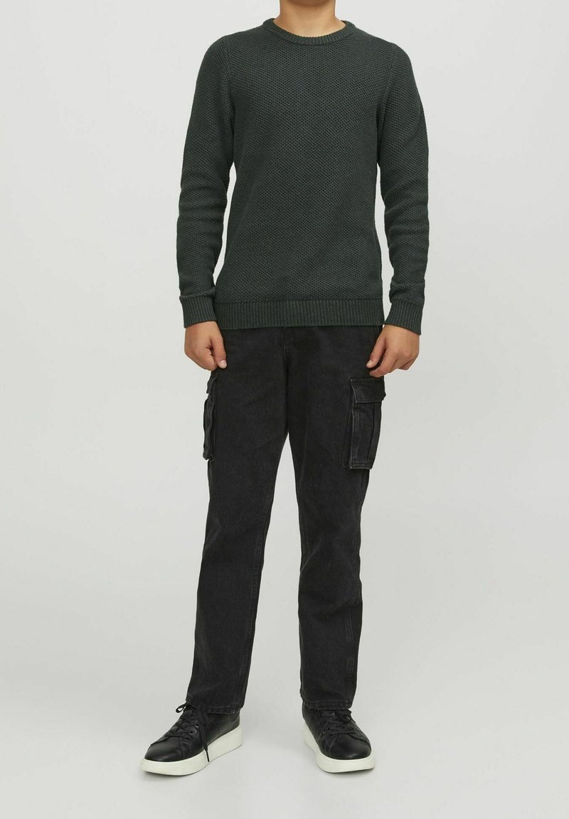 Barn i en mörkgrön texturerad tröja, svarta cargo-byxor med sidofickor och svarta sneakers med vita sulor, står mot en enfärgad bakgrund.