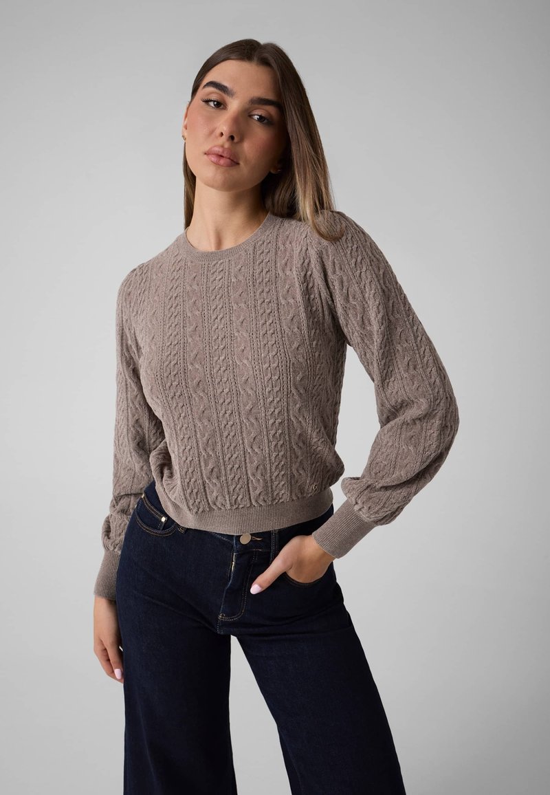 Femme aux longs cheveux bruns portant un pull en maille torsadée taupe et un jean bleu foncé, debout avec une main dans la poche, devant un fond gris.