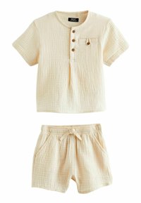 Svetlo béžová súprava loungewear z bavlny, ktorá obsahuje tričkovú košeľu s krátkym rukávom, trojicou gombíkov a vreckom, skombinovanú s šortkami na elastický pás.