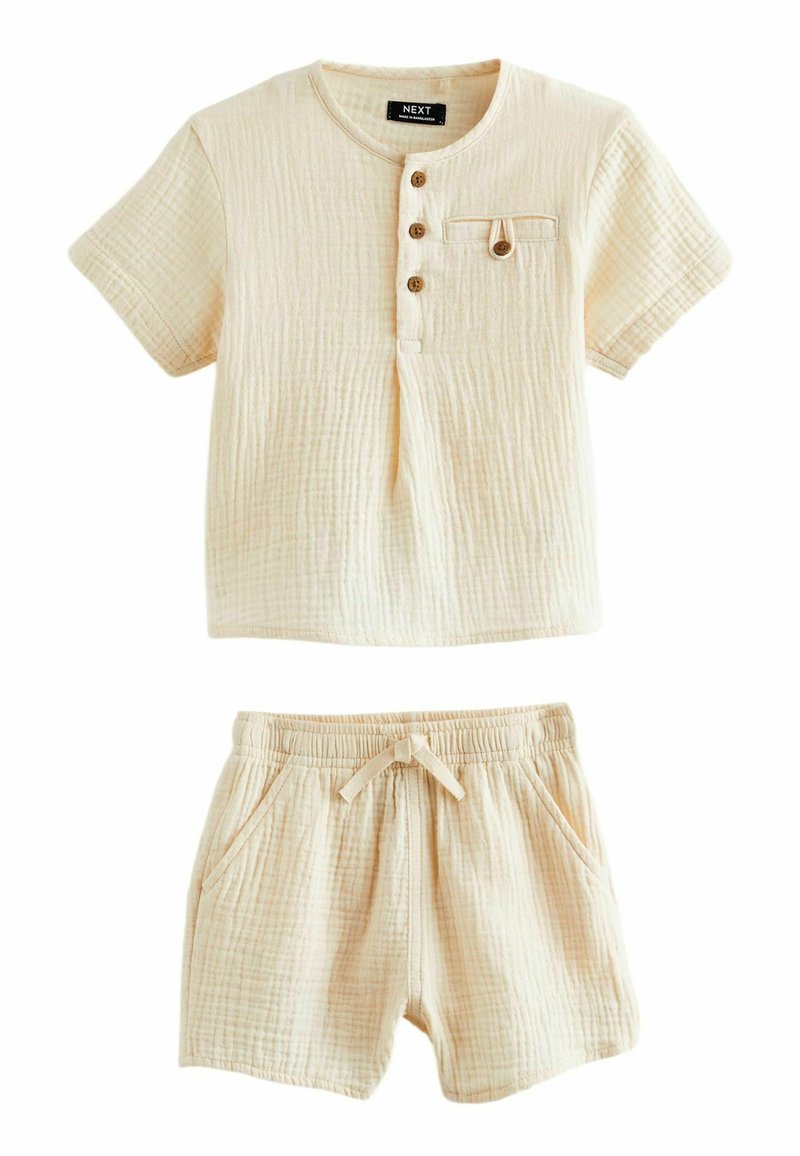 Svetlo béžová súprava loungewear z bavlny, ktorá obsahuje tričkovú košeľu s krátkym rukávom, trojicou gombíkov a vreckom, skombinovanú s šortkami na elastický pás.