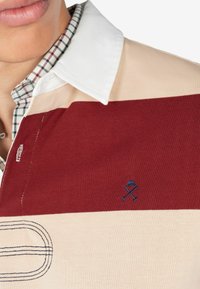 Polo a righe nei colori maroon e beige con colletto bianco. Dettagli del logo ricamati e fodera interna del colletto a quadretti.