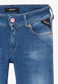 Jeans de mezclilla con un lavado azul desgastado, costuras en contraste y un botón rosa. La cinturilla incluye una etiqueta negra "REPLAY".