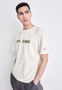 Beige bomull T-skjorte med korte ermer, med "PEPE JEANS" trykt i mørkebrun. Lite logo på venstre erme. Avslappet passform.