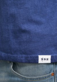 Granatowa bawełniana koszulka z miękką teksturą i małą białą metką na dole z szarymi akcentami. Częściowo wpuszczona w jasnoniebieskie dżinsy.