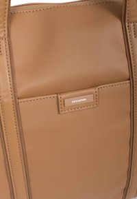 Tan totebag van gestructureerd synthetisch materiaal, met een voorzak en een rechthoekig label met de tekst "HEXAGONA" en zichtbare stiksels.
