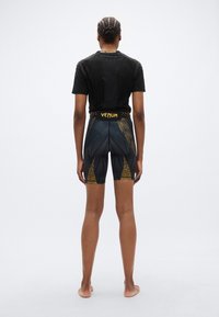 Chemise noire à manches courtes avec détails texturés et accents dorés, associée à un short noir moulant arborant un motif de lion doré et un logo.