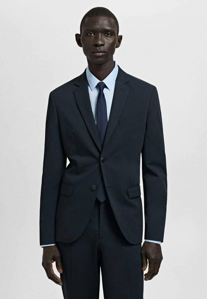 Donker marineblauw pak met een getailleerde pasvorm, een colbert met één knoop en bijpassende broek. Gecombineerd met een lichtblauw overhemd en een donkerstropdas.