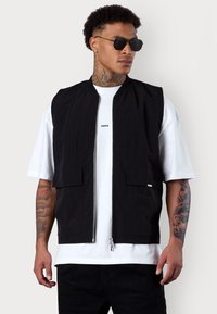 Schwarze ärmellose Jacke mit Stehkragen, Reißverschluss vorne und zwei frontalen Taschen, getragen über einem weißen T-Shirt, mit einer glatten Stofftextur.