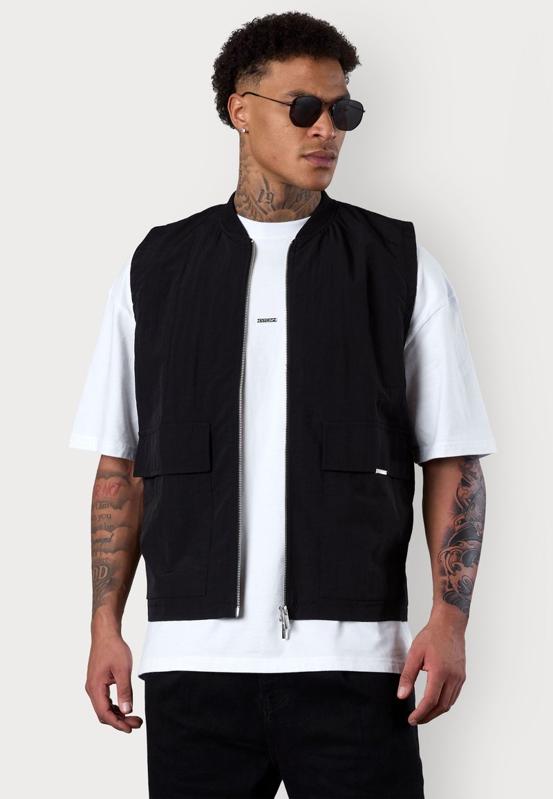 Schwarze ärmellose Jacke mit Stehkragen, Reißverschluss vorne und zwei frontalen Taschen, getragen über einem weißen T-Shirt, mit einer glatten Stofftextur.