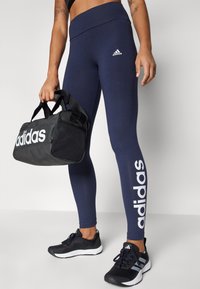 Jūrų jėgerių spalvos leggingai su aukštu juosmeniu ir baltu Adidas logotipu šone. Juodas sportinis krepšys su baltu Adidas tekstu ir tinklinėmis medžiagos detalėmis. Juodi sportbačiai.