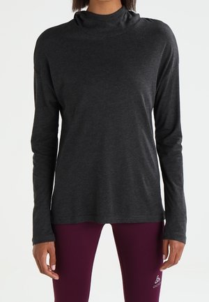 Personne portant un haut à manches longues gris foncé et un legging violet, debout devant un fond blanc uni.