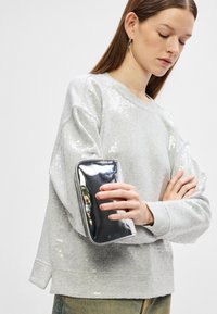 Mulher com um suéter de lantejoulas cinza claro, segurando uma bolsa de mão prateada brilhante, olhando para baixo com os braços cruzados sobre o peito.