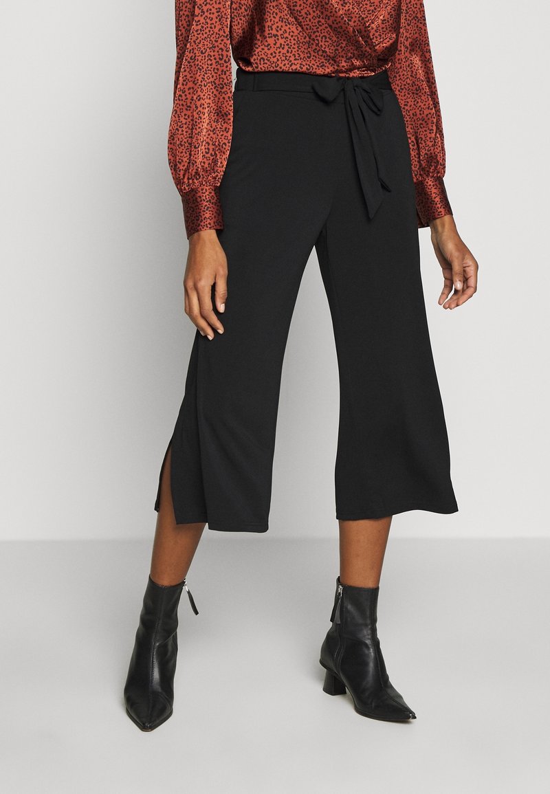 Femme portant un pantalon noir court à jambes larges avec des fentes sur les côtés, un chemisier rouge à motifs et des bottines noires avec des fermetures éclair.