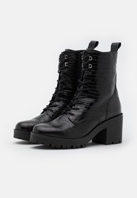 Bottines noires à texture crocodile, design à lacets, bout rond et talon épais. Dotées d'œillets métalliques et de languettes en boucle pour faciliter l'enfilage.