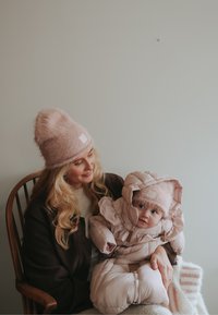 Vaaleanpunainen pörröinen pipo, jossa merkki, yhdistettynä vastaavaan tikattuun vauvan talvihaalariin. Pehmeät tekstuurit, pyöreät muodot ja hillityt vaaleanpunaiset yksityiskohdat.