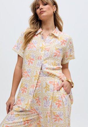 Frau trägt ein passendes kurzärmeliges Hemd und eine Hose mit einem abstrakten Blattmuster in Pastellorange, Gelb und Lila, goldene Creolen und Armbänder.