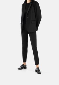 Blazer nero su misura con rever a punta, bottone singolo e tasche frontali, abbinato a pantaloni neri slim-fit e scarpe eleganti nere lucide.