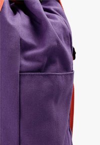 Gros plan sur la poche d'un sac à dos en tissu violet avec une sangle orange visible sur le côté, sur fond blanc.