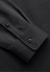 Poignet de chemise noire à manches longues avec une patte boutonnée, des bords cousus et une texture de tissu visible sur un fond noir.