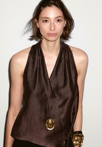 Top marrone senza maniche con design avvolto, realizzato in tessuto testurizzato. Caratterizzato da una lunga collana nera con un pendente dorato. Estetica semplice.