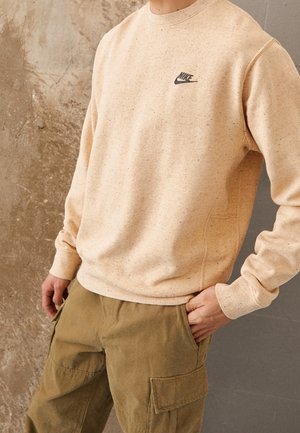 Sweatshirt beige clair en tissu texturé, avec un petit logo Nike noir sur la poitrine. Associé à un pantalon cargo vert olive.