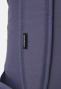 Mochila de tela morada con un acabado texturizado, correas acolchadas y una etiqueta de logo negra. Muestra parte del diseño lateral y de las correas.