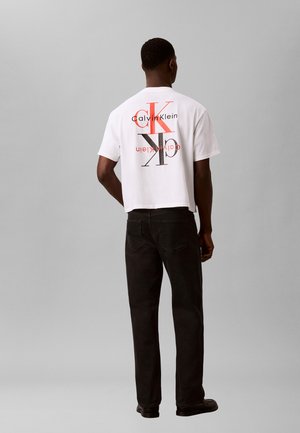 Mann, der mit dem Rücken zur Kamera steht, trägt ein weißes Calvin Klein T-Shirt mit großen orange-schwarzen CK-Logos und schwarze Hose vor grauem Hintergrund.