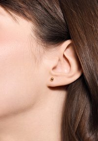 Złoty kolczyk wkręcany, mały, sferyczny kształt, gładka powierzchnia, umieszczony na płatku ucha osoby z prostymi brązowymi włosami.