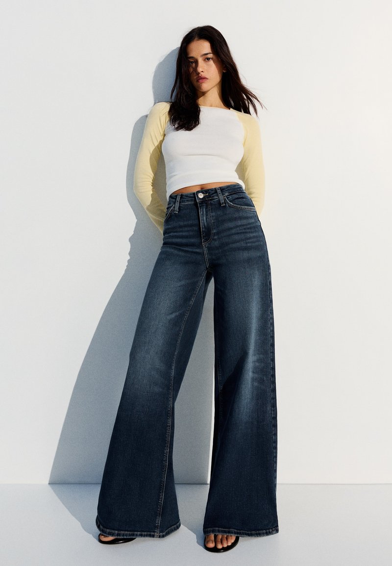 Jeune femme aux longs cheveux foncés portant un crop top blanc et jaune à manches longues et un jean large taille haute foncé, debout contre un mur blanc.