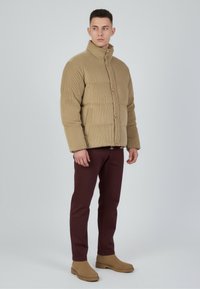 Kurtka puchowa w kolorze khaki z teksturowaną powierzchnią, posiadająca wysoki kołnierz, zapięcia na guziki oraz dwie boczne kieszenie, zestawiona z ciemnymi bordowymi spodniami i beżowymi botkami.