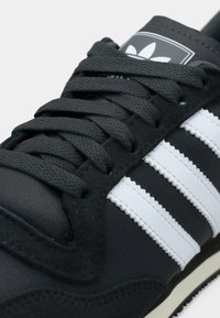 Black sneaker with white stripes, black laces, and Adidas trefoil logo on the tongue, shown in close-up.