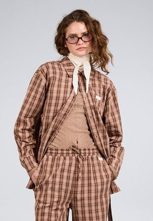 Junge Person mit Brille, braun kariertem Hemd und Hose, hellbraun strukturiertem Oberteil und einem weißen Schal um den Hals gebunden, Hände in den Taschen.