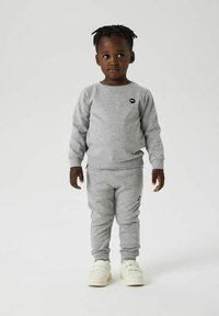 Grå hettegenser og bukse sett laget av mykt stoff. Har en liten svart logo på brystet og detaljer med side lommer. Hvite sneakers.