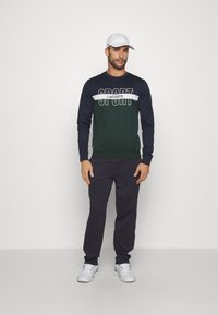 Sudadera azul marino y verde con "LACOSTE SPORT" en blanco, combinada con pantalones azul marino y zapatillas blancas, bajo una gorra blanca.