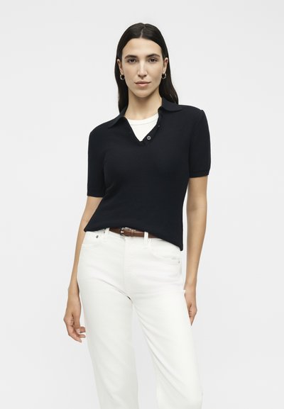 Γυναίκα με μακριά σκούρα μαλλιά φοράει μαύρο πόλο με κοντά μανίκια πάνω από λευκό τοπ, λευκό παντελόνι και καφέ ζώνη μπροστά σε μονόχρωμο φόντο.