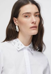 Chemise blanche avec un col orné de perles et d'ornements, comportant des accents argentés. Le tissu semble lisse avec une texture subtile. Les boucles d'oreilles sont métalliques et de forme ovale.