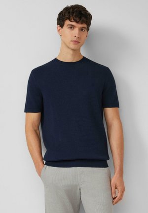 Jeune homme aux cheveux bouclés portant un pull à manches courtes bleu marine et un pantalon gris clair, debout avec une main dans la poche sur un fond uni.