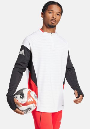 Langarm sporttop in het wit met zwarte en rode accenten, voorzien van een ritssluiting in de nek en duimgaten, gecombineerd met een patroonvoetbal.