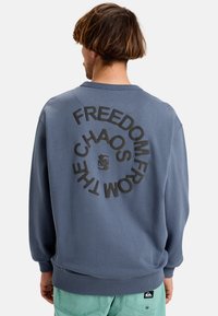 Felpa blu con scollo rotondo e polsini a costine, caratterizzata da un design grafico circolare di testo nero sulla schiena e un'etichetta con logo sull'orlo.