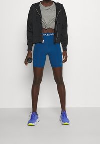 Černá mikina na zip, šedé uvázané tričko, modré šortky a fialové sportovní boty se žlutými detaily. Drží malé černé činky.