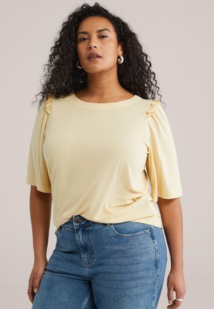 Donna con capelli ricci indossa una maglietta gialla chiaro a maniche corte con spalle arricciate e jeans a vita alta blu, in piedi davanti a uno sfondo semplice.